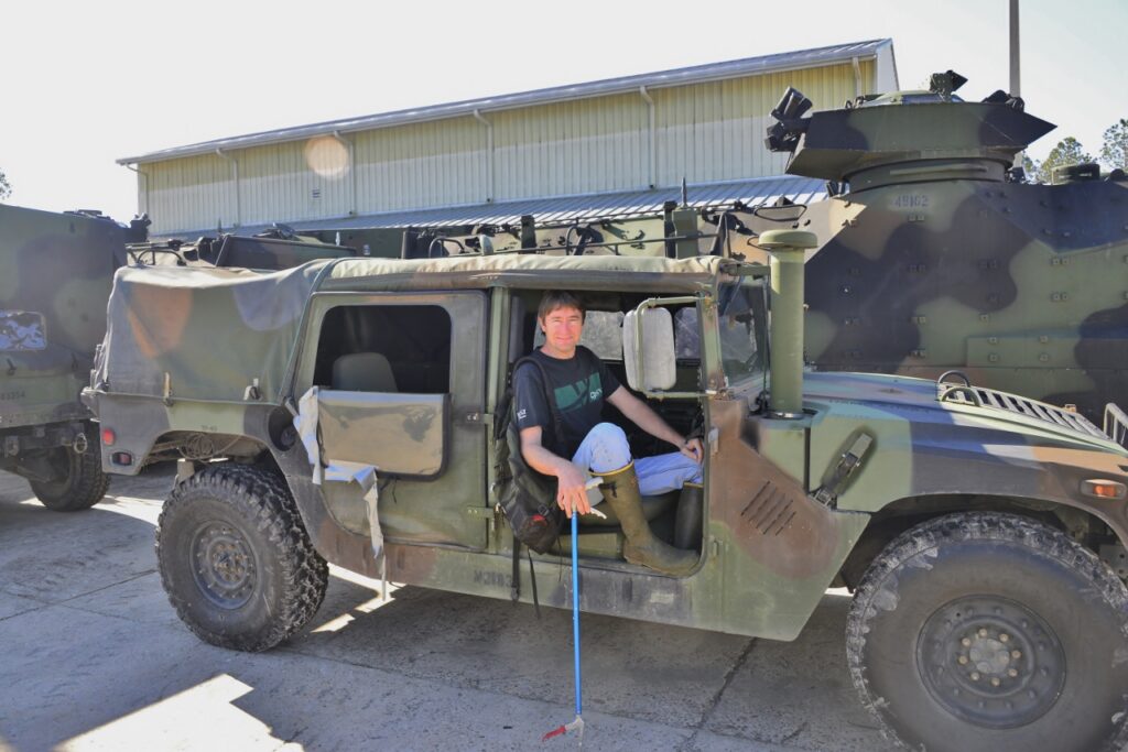 Chris sits in a military vehicle in a black shirt with knee boots and a snake hook. 